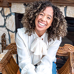 Eczema advocate Ashley Ann Lora flashing a huge smile in her seat in front of a stone fireplace.