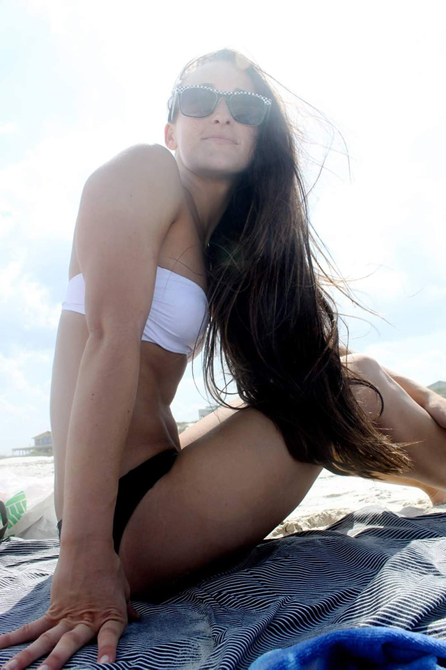 Briana Banos wearing sunglasses and a swimsuit, sitting on a beach.